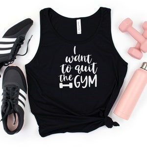 Puede incluir: Camiseta de tirantes negra con el texto "I want to quit the gym" en letras blancas con un icono de mancuerna. La camiseta está sobre una superficie blanca con un par de zapatillas negras, una botella de agua rosa y mancuernas rosas.