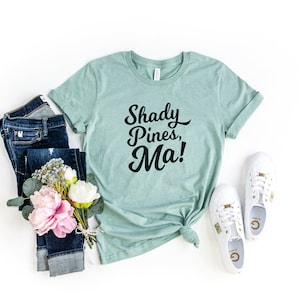 May include: A teal blue t-shirt with the text "Shady Pines, Ma!" in black script font. The shirt is paired with blue jeans and white sneakers.