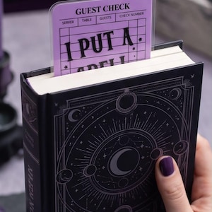 May include: A black book with a celestial cover design featuring moons and stars, containing a light purple bookmark shaped like a guest check. The bookmark displays the text GUEST CHECK at the top and the words I PUT A SPELL on the lower section.