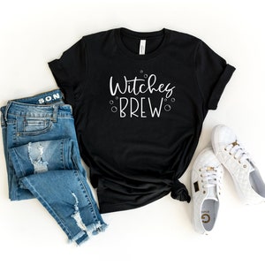 Op de afbeelding: Zwart T-shirt met de tekst "Witches Brew" in witte letters. Het shirt wordt gecombineerd met een blauwe jeans en witte sneakers.
