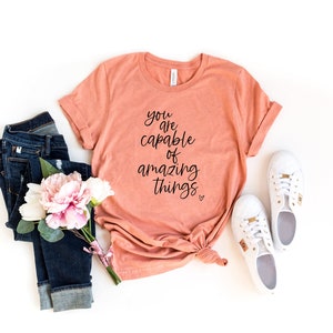 Può includere: Una maglietta color pesca con il testo "you are capable of amazing things" in corsivo nero. La maglietta è stesa su uno sfondo bianco con un paio di jeans blu, un mazzo di fiori rosa e scarpe da ginnastica bianche.