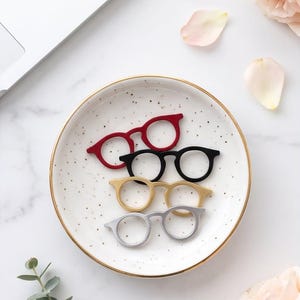 May include: A set of four rings shaped like eyeglasses in red, black, gold, and silver tones. The rings are arranged in a stack on a round white ceramic dish with gold trim and small brown speckles, featuring circular frames and thin bands.