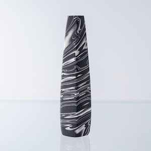 Puede incluir: Un jarrón alto y esbelto con un patrón jaspeado de remolinos blancos y negros. El objeto tiene una forma geométrica cónica con una abertura superior plana y un acabado mate suave que se extiende desde el cuello estrecho hasta la base.