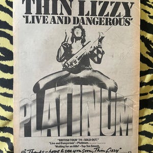 May include: A vintage black and white print advertisement for the band Thin Lizzy featuring the text THIN LIZZY LIVE AND DANGEROUS and PLATINUM. The graphic shows a musician playing a guitar, with additional text detailing a 1979 British tour.
