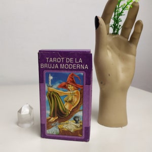 May include: A purple box with the text "Tarot de la Bruja Moderna" on the front. The box contains a tarot deck with a colorful illustration of a woman in a witch's hat holding a sword.