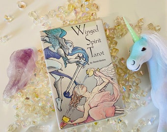 May include: A tarot deck box with the title "Winged Spirit Tarot" by David Sexton. The box is surrounded by iridescent crystals and a blue unicorn toy.