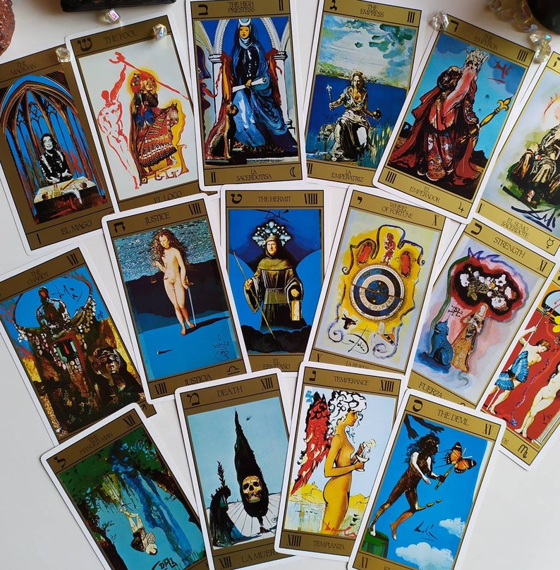 Puede incluir: Un conjunto de cartas de tarot con ilustraciones coloridas y detalles dorados. Las cartas presentan varios s&iacute;mbolos y personajes, incluyendo un mago, un ermita&ntilde;o y un diablo. Las cartas est&aacute;n dispuestas en semic&iacute;rculo sobre una superficie blanca.