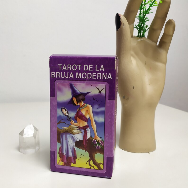 May include: A purple box with the text "Tarot de la Bruja Moderna" on the front. The box features an illustration of a woman in a witch's hat and dress, holding a magnifying glass and a book.