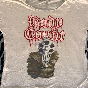 May include: Off-white t-shirt featuring the text "Body Count" in red gothic lettering with dripping blood effect. Below the text is a graphic of a hand holding a revolver. The shirt has a crew neck.