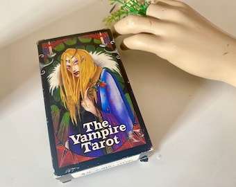 May include: A box for The Vampire Tarot deck. The box features a colorful illustration of a woman with long blonde hair, wearing a purple and blue dress, holding a red rose. The background is a dark green and black forest scene.
