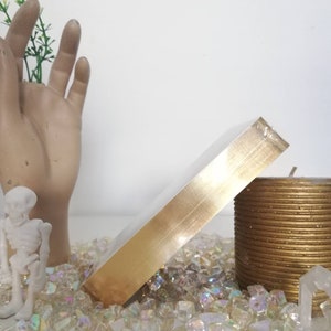 Puede incluir: Una caja de color dorado con un acabado met&aacute;lico brillante. La caja est&aacute; sentada sobre un lecho de piedras de vidrio iridiscentes y junto a una vela dorada.