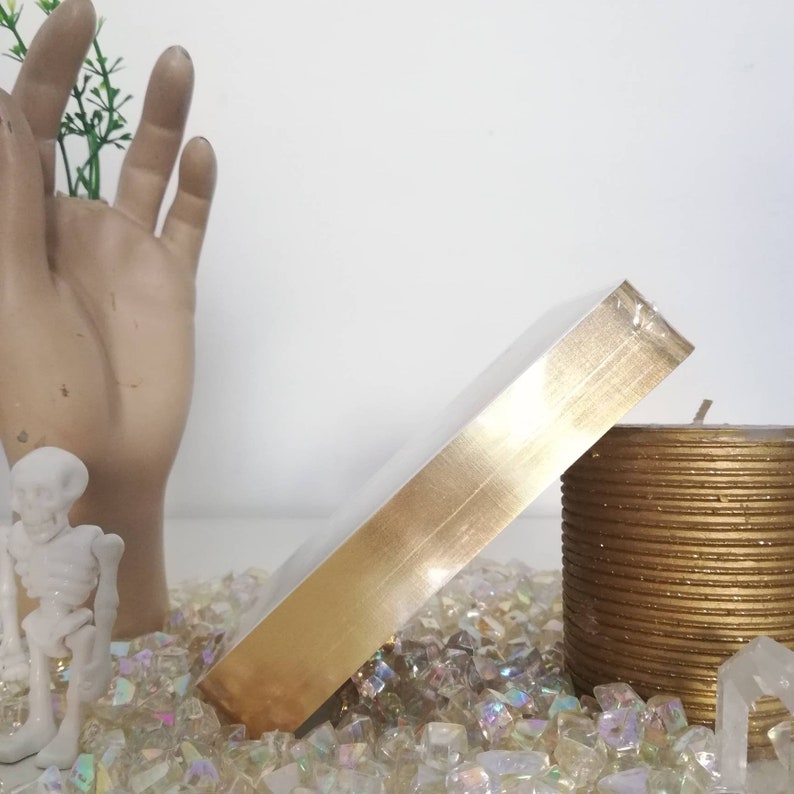 Puede incluir: Una caja de color dorado con un acabado met&aacute;lico brillante. La caja est&aacute; sentada sobre un lecho de piedras de vidrio iridiscentes y junto a una vela dorada.