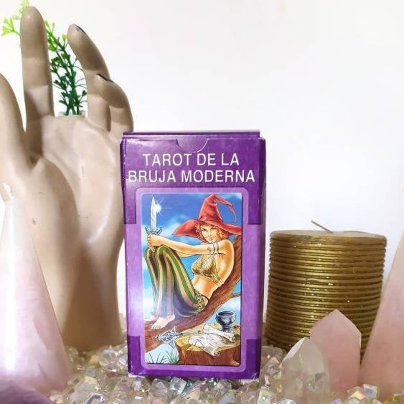 May include: Tarot deck box with a purple background and the text "Tarot de la Bruja Moderna". The box features an illustration of a woman with a sword and a crown, sitting on a tree branch.