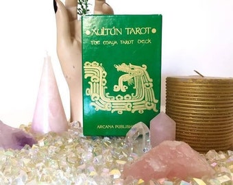 Puede incluir: Una caja rectangular verde de cartas de tarot titulada Xultún Tarot The Maya Tarot Deck con letras doradas y una ilustración de serpiente dorada. La caja está colocada cerca de una mano de maniquí beige, una vela cónica rosa y varios cristales.