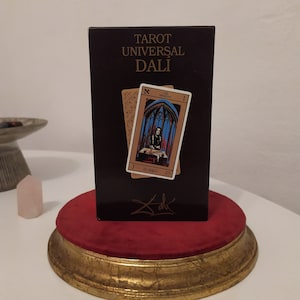 Puede incluir: Una caja negra con letras doradas que dicen "Tarot Universal Dal&iacute;" y una firma de Salvador Dal&iacute;. La caja tiene dos cartas de tarot en el frente, una con una mujer con un vestido azul y rojo y la otra con un fondo dorado.