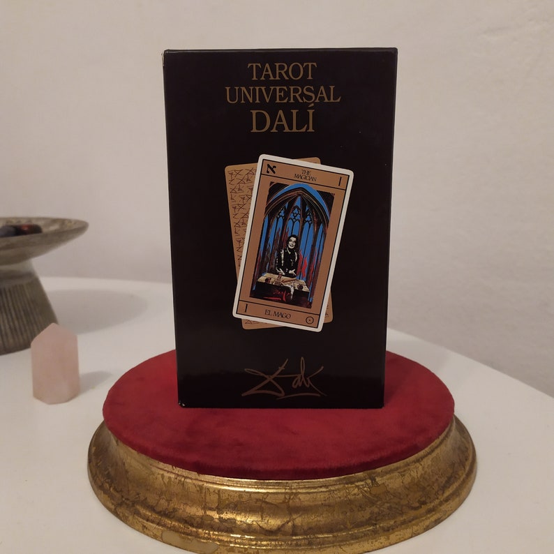 Puede incluir: Una caja negra con letras doradas que dicen "Tarot Universal Dal&iacute;" y una firma de Salvador Dal&iacute;. La caja tiene dos cartas de tarot en el frente, una con una mujer con un vestido azul y rojo y la otra con un fondo dorado.