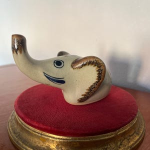 Puede incluir: Una figurita de elefante de cerámica con detalles pintados en marrón y azul, sentada sobre una base de terciopelo rojo con ribete dorado.