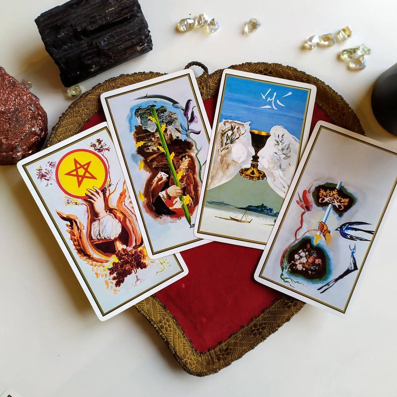 Puede incluir: Cuatro cartas de tarot sobre un fondo de terciopelo rojo. Las cartas presentan ilustraciones de un pentagrama, un c&aacute;liz, una guada&ntilde;a y un p&aacute;jaro. Las cartas est&aacute;n rodeadas de cristales.