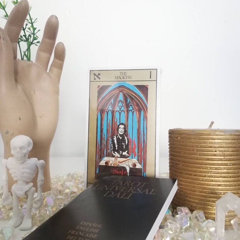 Puede incluir: Baraja de tarot con una ilustraci&oacute;n colorida de la carta El Mago de la baraja Tarot Universal Dali. La carta muestra a un hombre con una t&uacute;nica roja y negra, barba y bigote blancos, sentado a una mesa con un mantel blanco. La carta est&aacute; rodeada de un marco dorado con las palabras "El Mago" e "I" en texto negro.