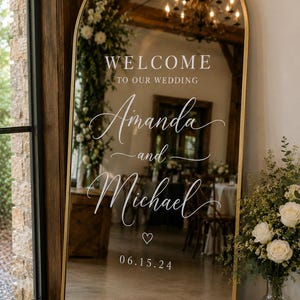 May include: An arched floor mirror with a thin gold frame featuring white text that reads WELCOME TO OUR WEDDING, Amanda and Michael, and the date 06.15.24 with a small heart icon. The mirror reflects an indoor event space with a chandelier.