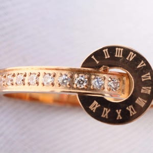 Könnte beinhalten: Ein goldfarbener Ring mit einem Band, das mit kleinen, klaren Edelsteinen besetzt ist. Ein kreisförmiger Anhänger mit römischen Ziffern ist am Band befestigt. Der Ring ist vor einem weichen, neutralen Hintergrund platziert.