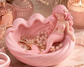 May include: A pink seashell-shaped trinket dish with a mermaid figurine. The mermaid has pink hair and a tail, and is resting in the dish, which holds jewelry and pearls. A lit candle and perfume bottles are in the background.
