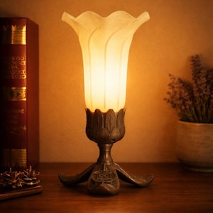 May include: A table lamp featuring a frosted white glass shade shaped like a flower blossom, set into a dark metallic base with leaf-like details. The base includes a small circular emblem with the text "GG MOON" embossed on the front.