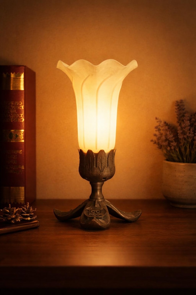 May include: A table lamp featuring a frosted white glass shade shaped like a flower blossom, set into a dark metallic base with leaf-like details. The base includes a small circular emblem with the text "GG MOON" embossed on the front.