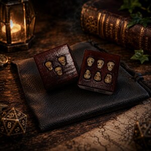 May include: Two dark brown square dice featuring small, off-white skull carvings embedded on their faces. One die displays three skulls, while the other die displays six skulls. The dice have a polished, wood-like finish and are resting on a black pouch.