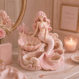 Könnte beinhalten: Eine rosa Meerjungfrau-Figur mit langen Haaren und einem Muschel-BH sitzt auf einem dekorativen Schmuckhalter. Der Halter hat wellenförmige Designs und muschelförmige Fächer, perfekt für die Aufbewahrung kleiner Schmuckstücke. Ein gerahmter Druck mit den Worten "You are so loved" ist sichtbar.