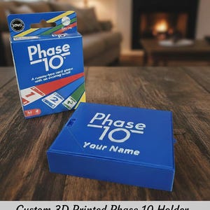 Op de afbeelding: Een blauwe Phase 10 kaartspeldoos en een bijpassende houder worden getoond op een houten oppervlak. De doos heeft de speltitel in het wit en de houder heeft ruimte voor een naam. De speldoos is blauw met witte tekst.