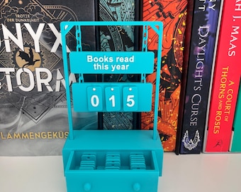 May include: A teal book counter with the text "Books read this year" and the number "015" displayed. The counter has a drawer at the bottom and is set against a backdrop of colorful books on a shelf.