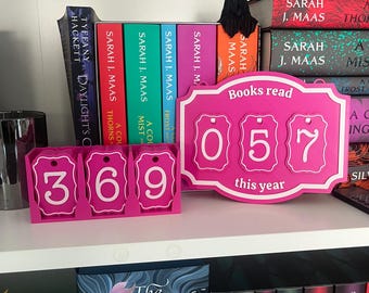 May include: A shelf displays books and a pink counter for tracking reading progress. The counter reads "Books read 057 this year" and has additional number tags showing "369". The books have colorful spines and include titles by Sarah J. Maas.