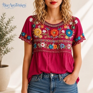 Peut inclure: Un chemisier magenta à manches courtes orné de broderies florales multicolores complexes sur la poitrine et les manches, avec un ourlet festonné sur les manches et une encolure ronde. Le texte Blue Aura Trinkets apparaît en haut à gauche.