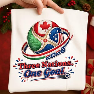 May include: White t-shirt with a soccer ball design featuring the flags of Canada, the United States, and Mexico. The design includes the text "2026" and "Three Nations. One Goal."