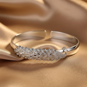 Könnte beinhalten: Ein silberfarbenes Armband mit Pfauenfedermuster. Das Armband hat ein offenes Manschettendesign und ist mit filigranen Details und kleinen, funkelnden Verzierungen versehen. Es wird auf einer goldenen Satin-Oberfläche präsentiert.