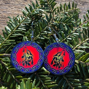 May include: A pair of silver hoop earrings with a red, orange, and blue wolf design. The wolf is howling and is surrounded by a blue Celtic knot design.
