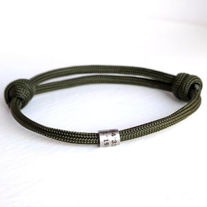 May include: An olive green cord bracelet featuring a cylindrical silver-toned metal bead engraved with the coordinates 54 31 and 1 19. The bracelet is constructed from a woven cord with two adjustable sliding knots on either side of the band.