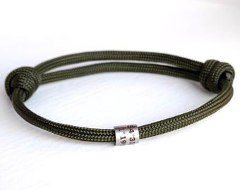 May include: An olive green cord bracelet featuring a cylindrical silver-toned metal bead engraved with the coordinates 54° 31' and 1° 19'. The bracelet is secured with two adjustable sliding knots on either side of the central bead.