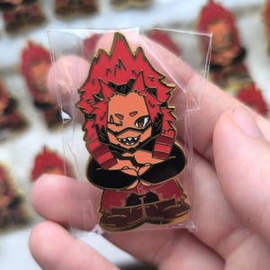 Pode incluir: Um pin de esmalte vermelho e preto com um personagem de cabelo vermelho espetado e uma expressão determinada. O pin está em uma manga de plástico transparente, com vários pinos semelhantes visíveis no fundo.