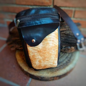 Puede incluir: Riñonera de cuero negro con un panel frontal texturizado de color marrón claro. La bolsa tiene una cremallera en la parte superior, una solapa con un broche plateado y una correa ajustable marrón. La bolsa está sobre una superficie de madera.