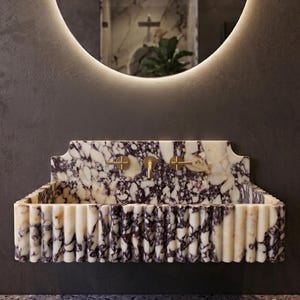 May include: A marble bathroom sink with a fluted design and gold fixtures. The sink has a white and purple veined pattern. The sink is mounted on a dark grey wall, with a round mirror above it.