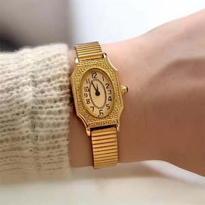 May include: A gold-toned wristwatch with an oval face and a stretch band. The watch face features black numerals and hands against a cream-colored background. The watch is worn on a wrist.