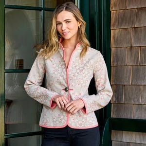May include: A beige jacket with a floral pattern and pink trim. The jacket has a mandarin collar, long sleeves, and a single dark button closure. The floral design is in shades of pink and beige. The model is wearing black jeans.