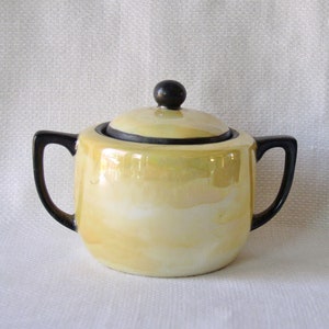 Puede incluir: Un azucarero amarillo con tapa negra y asas negras. El tazón tiene un acabado perlado.