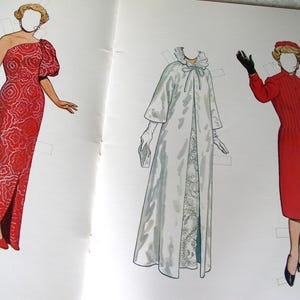 May include: A paper doll set featuring three figures with blank faces. The left figure wears a red one-shoulder gown with a white floral pattern. The center figure wears a long white coat with a ruffled collar. The right figure wears a red coat.
