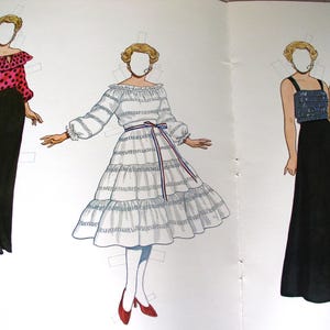 May include: A paper doll set featuring three figures with blonde hair and blank faces. The first wears a pink polka-dot blouse and black skirt. The second wears a white tiered dress with a red, white, and blue sash. The third wears a blue sequined top and black skirt.