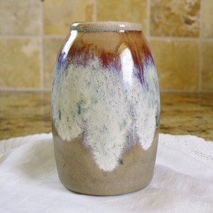 May include: A small, round ceramic vase with a brown, white, and green glaze. The vase has a mottled, textured surface and a slightly flared lip.