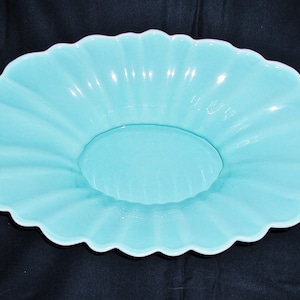 May include: A light blue, oval-shaped serving dish with a scalloped edge. The dish has a central, slightly raised area and a fluted design radiating from the center to the rim. The color is a soft, pastel blue.