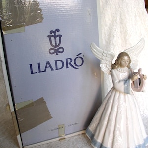May include: A porcelain angel figurine playing a small harp, featuring a long white dress with blue trim and detailed wings, positioned next to a light blue cardboard box with the Lladro logo and the text Valencia España printed on the front.
